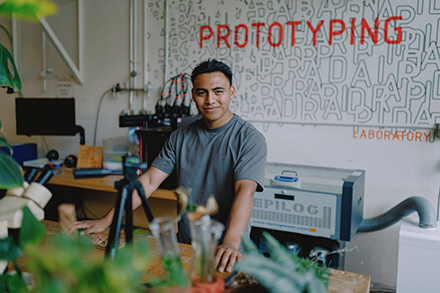 An SFSU student smiles in a plant-filled classroom with a sign reading ‘Prototyping Laboratory’ behind them.